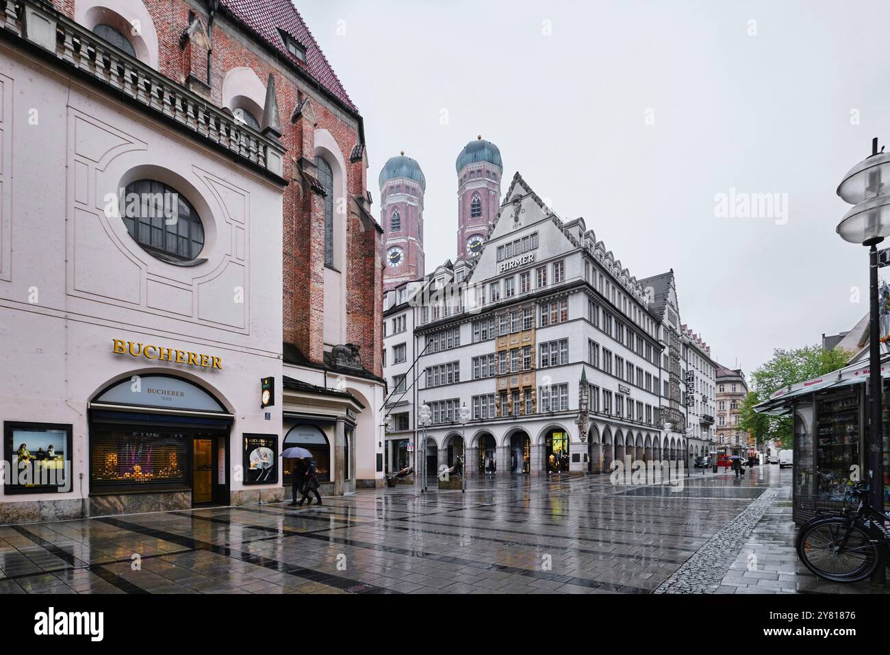 München - 18. April 2024: Außenansicht des größten Herrenmode-Hauses Hirmer. Die Türme der Frauenkirche Stockfoto