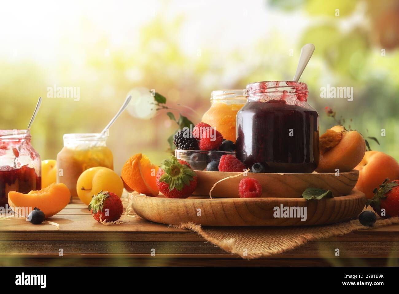 Mehrere Gläser mit Marmelade verschiedener Geschmacksrichtungen mit verschiedenen Früchten auf einem rustikalen Holztisch mit einem Hintergrund von Feldfrüchten in der Natur. Vorderansicht. Stockfoto