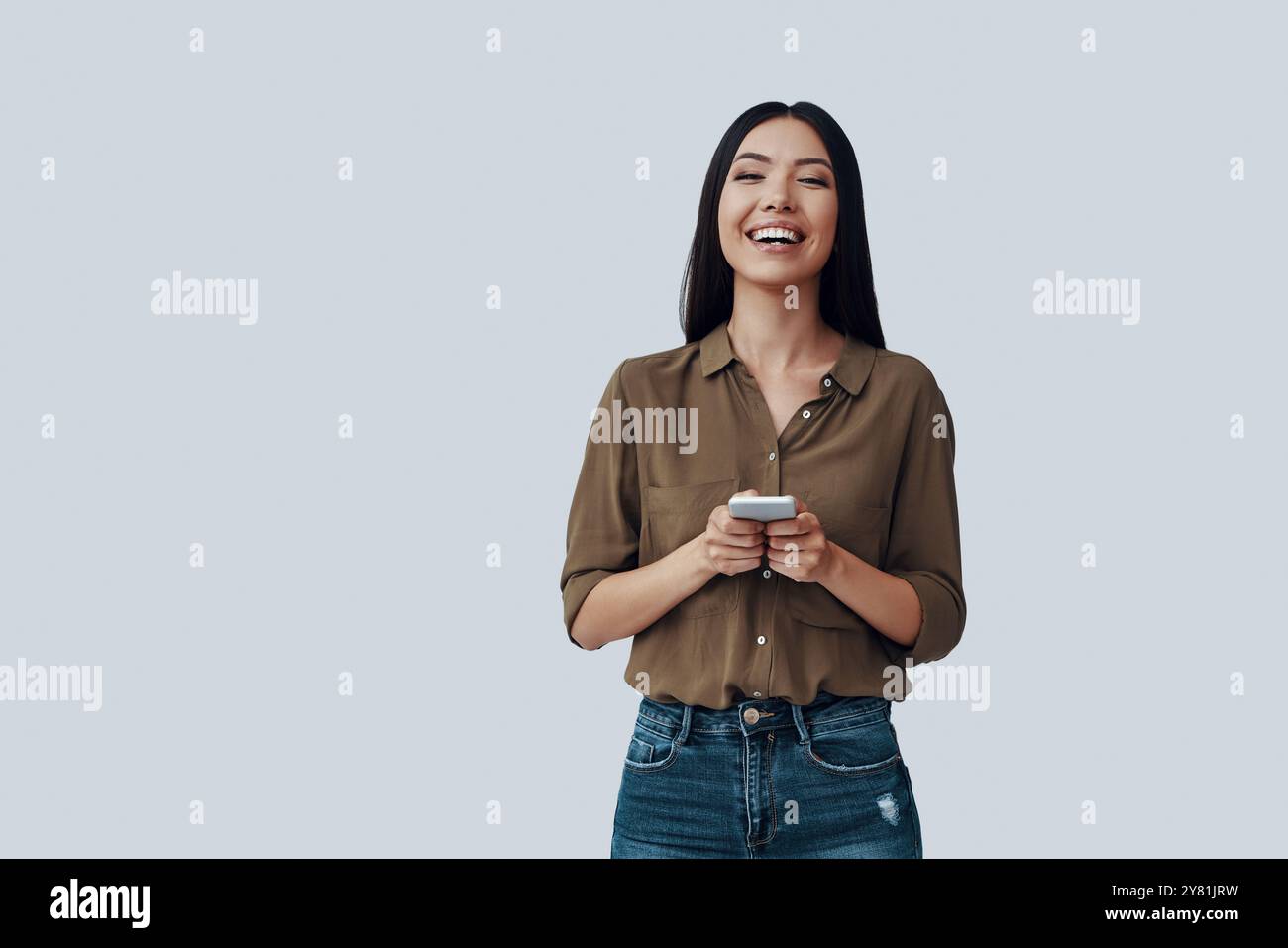 Gute Nachrichten empfangen. Attraktive junge asiatische Frau, die ihr Smartphone benutzt und lächelt, während sie vor grauem Hintergrund steht Stockfoto