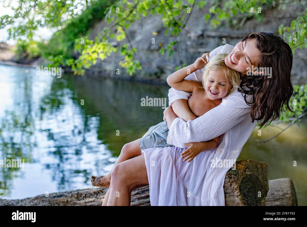 Eine fröhliche Frau umarmt einen kleinen Jungen an einem ruhigen Seeufer, der sowohl lächelt als auch einen sonnigen Tag im Freien genießt. Stockfoto