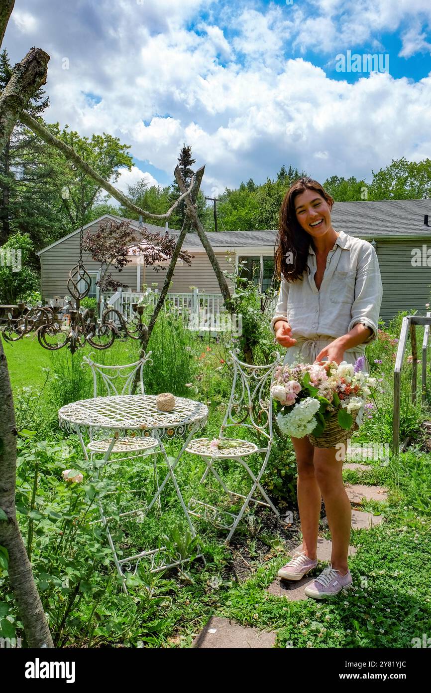 Lächelnde Frau, die in einem üppigen Garten steht und einen Blumenstrauß neben alten Gartenmöbeln unter einem klaren, sonnigen Himmel arrangiert. Stockfoto
