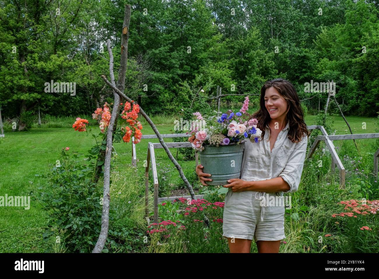 Lächelnde Frau, die in einem üppigen Garten steht und einen Eimer voller bunter Blumen hält. Stockfoto