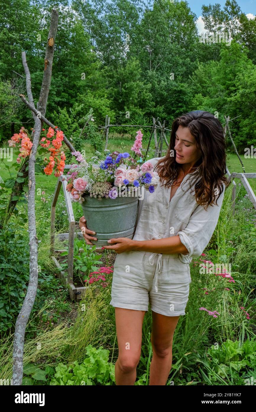 Eine Frau, die einen Eimer voller bunter Blumen in einem üppigen Garten hält. Stockfoto