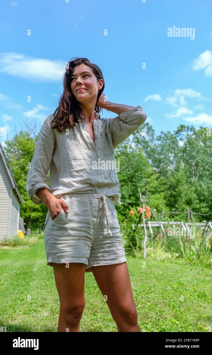 Frau, die an einem sonnigen Tag in einem Garten steht, ihre Hand auf der Hüfte und die andere berührte ihren Hals und lächelte zum Himmel. Stockfoto