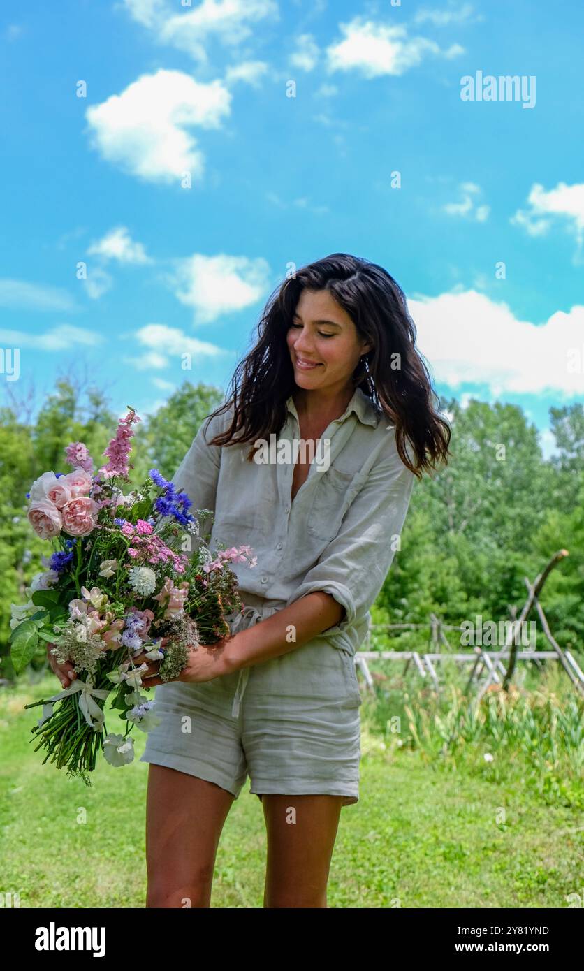 Eine Frau in lässigem Outfit lächelt, während sie einen Blumenstrauß auf einem sonnigen Feld hält. Stockfoto
