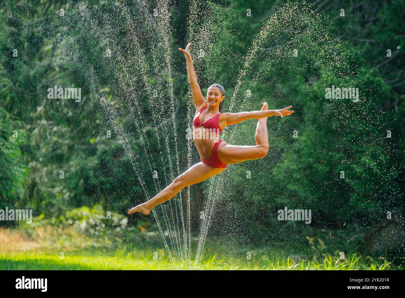 Eine freudige Frau führt einen Ballettsprung zwischen Wassertropfen auf einem grasbewachsenen Rasen durch. Stockfoto