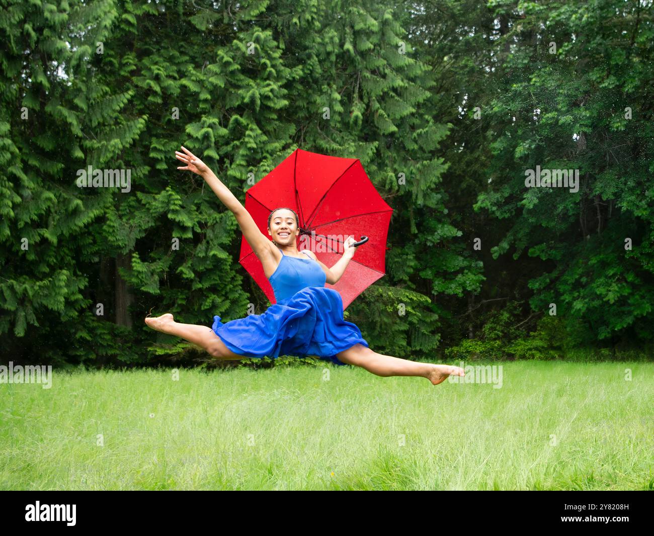 Fröhliche Frau in blauem Kleid tanzt auf einem grünen Rasen mit einem roten Regenschirm. Stockfoto