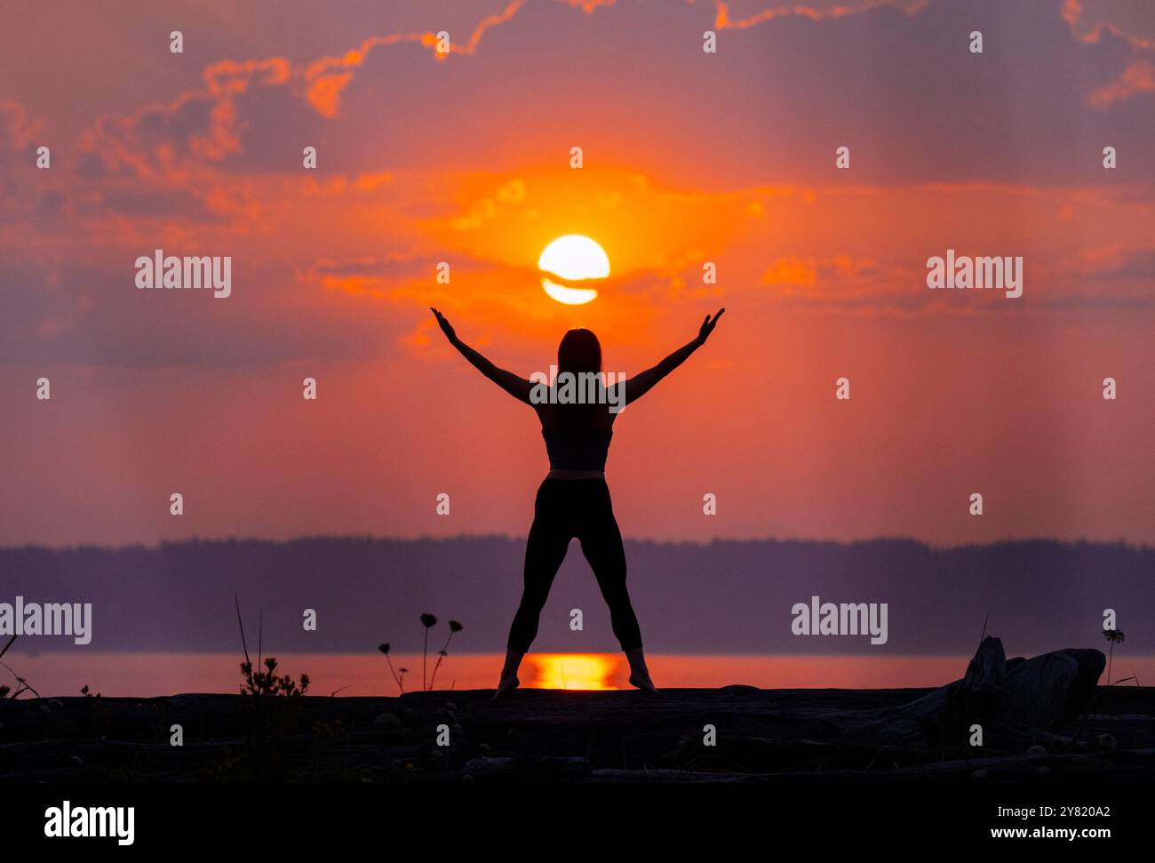 Silhouette einer erwachsenen Frau mit erhobenen Armen, die vor einem lebendigen Sonnenuntergang über einem ruhigen See steht. Stockfoto