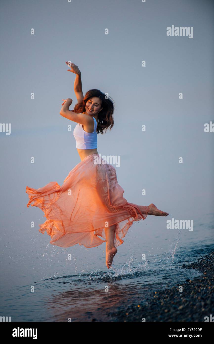 Fröhliche Frau tanzt in der Dämmerung am Strand mit einem fließenden pfirsichfarbenen Kleid. Stockfoto