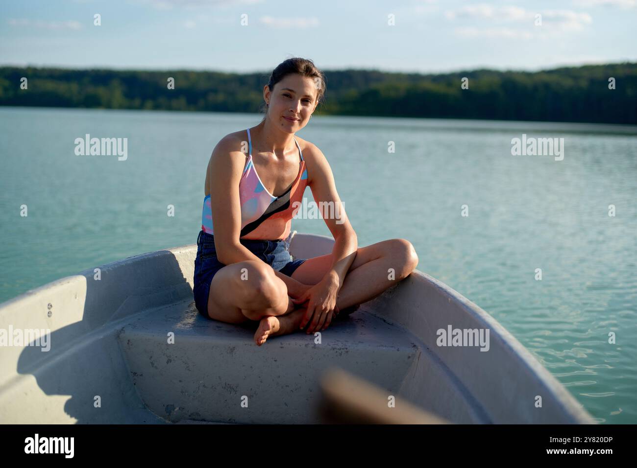 Junge Frau lächelt, während sie im Kreuz auf dem Bug eines Bootes auf einem ruhigen See sitzt. Stockfoto