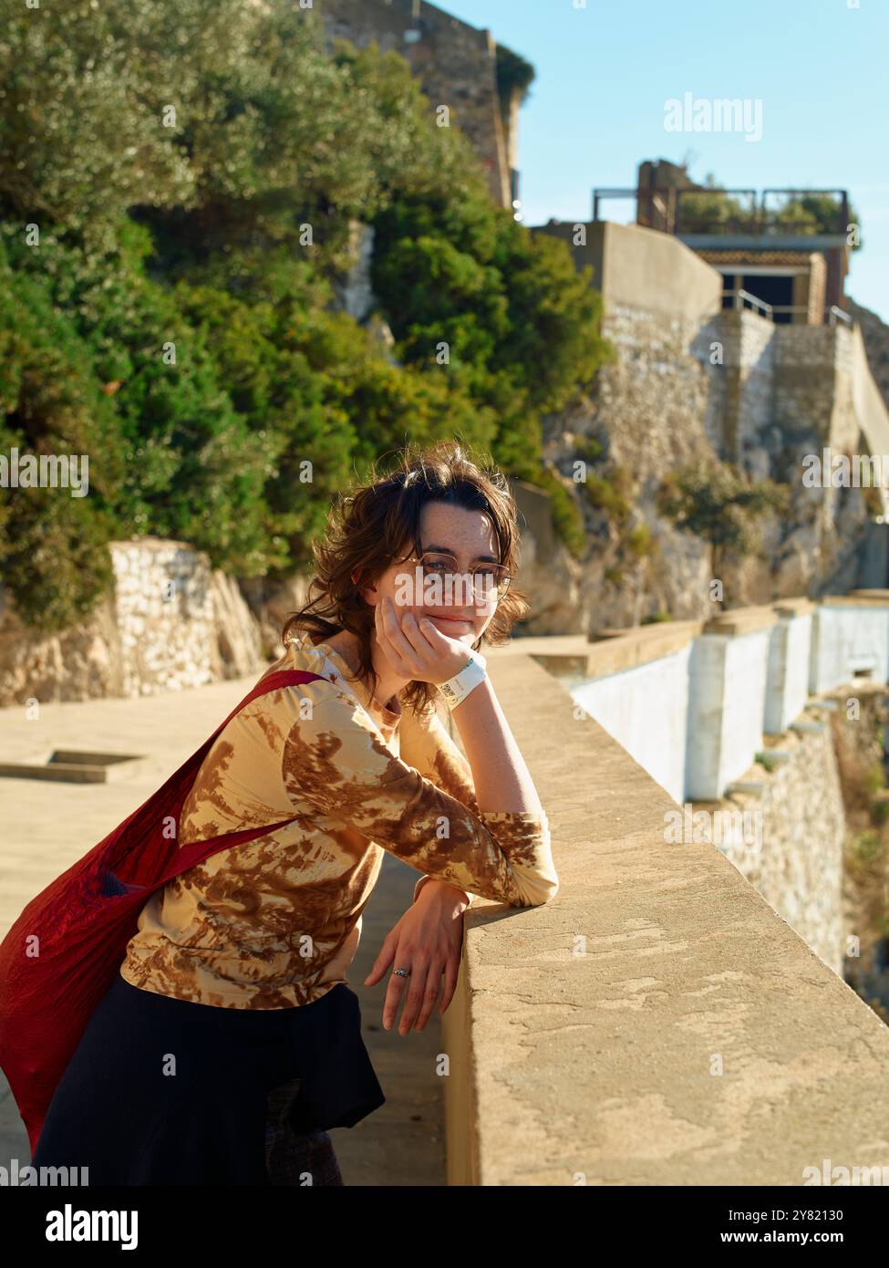 Lächelnde junge Frau, die sich an einem sonnigen Tag auf einem Steingeländer mit einem historischen Gebäude im Hintergrund lehnt. Stockfoto