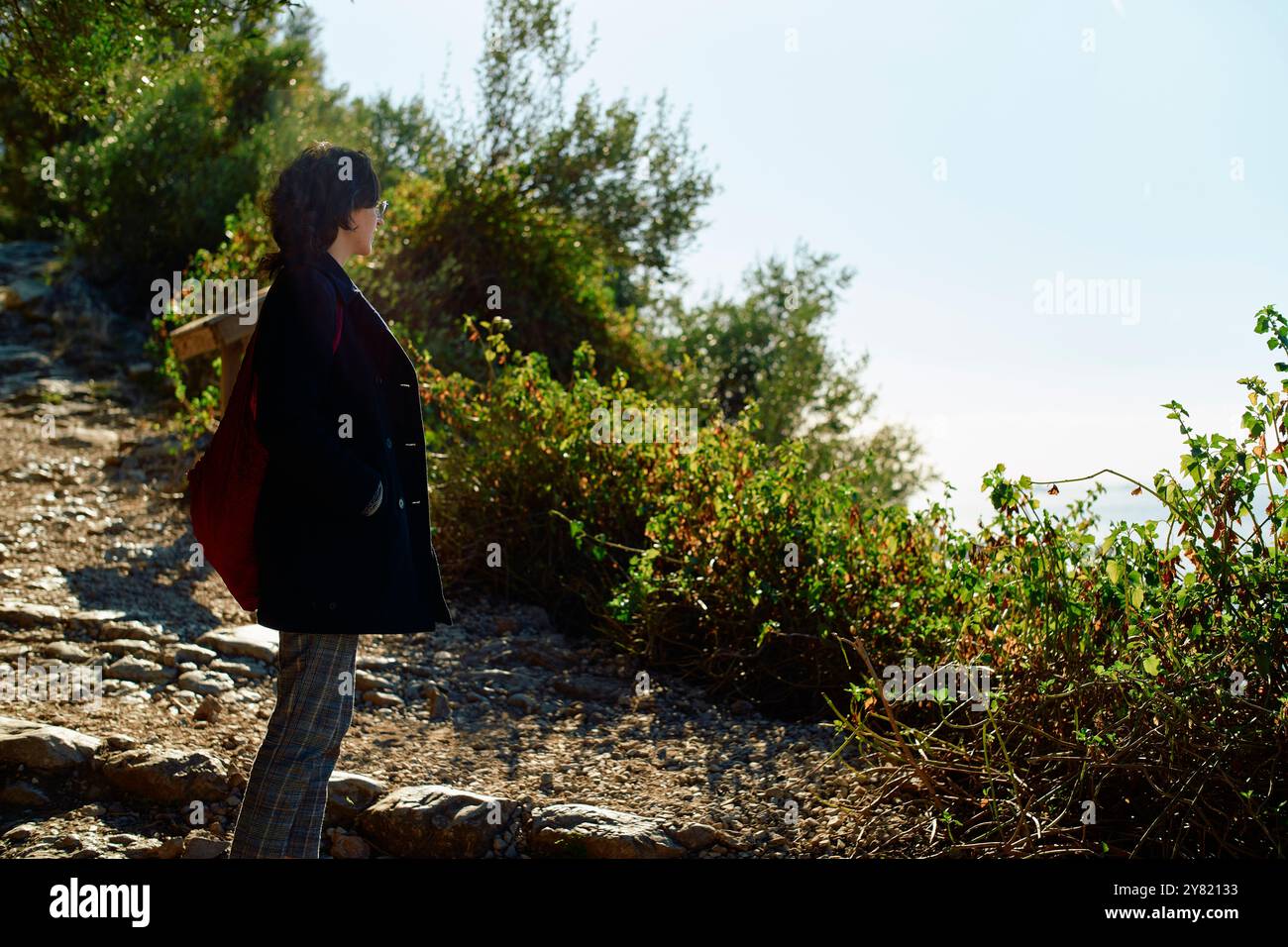 Frau mit roter Tasche geht auf einem unbefestigten Weg, umgeben von grünem Sträucher, durch den das Sonnenlicht gefiltert wird. Stockfoto