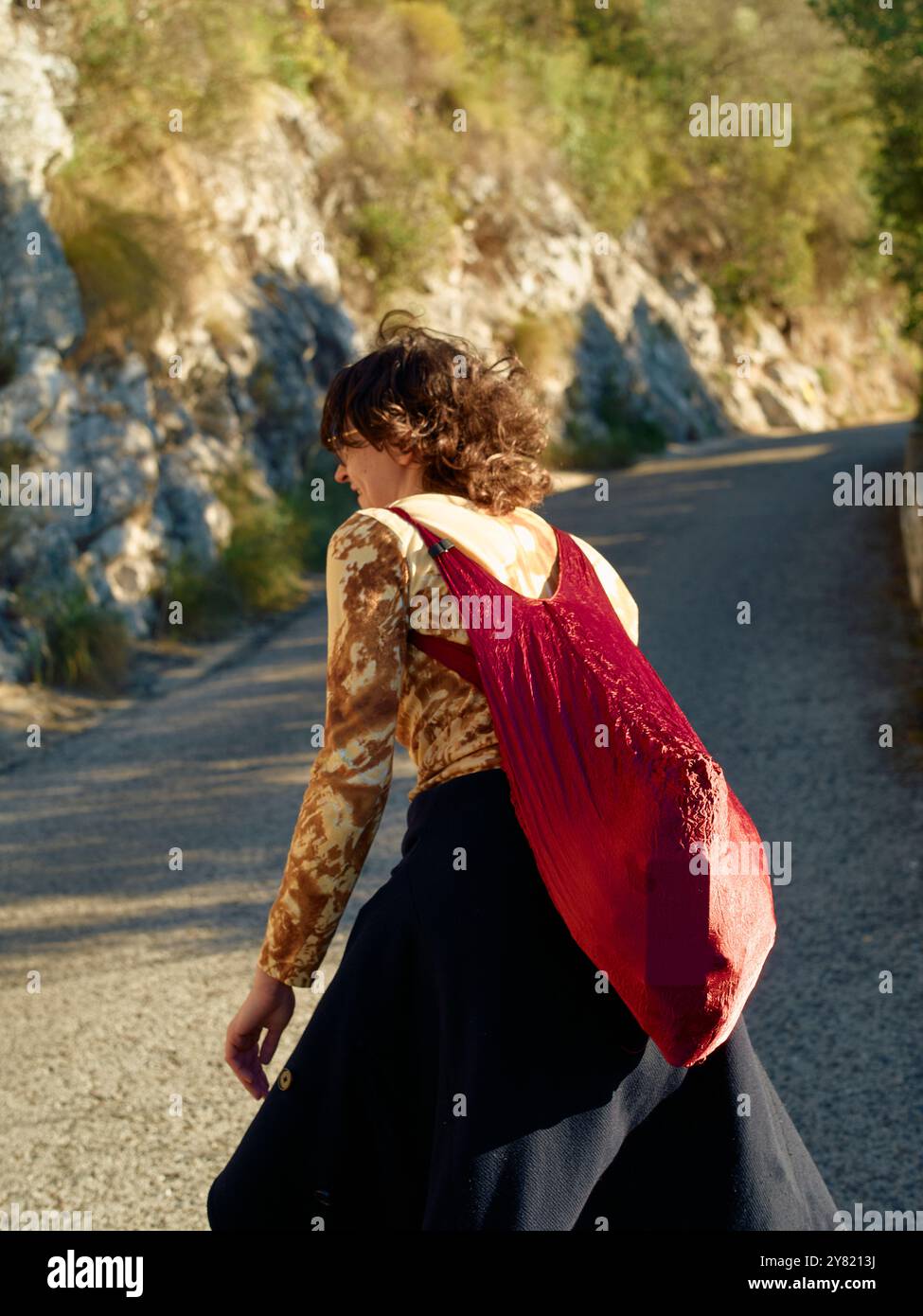Eine Frau geht auf einer malerischen Straße mit einer roten Tasche, gekleidet in einem einzigartigen gemusterten Oberteil und einem schwarzen Rock. Stockfoto