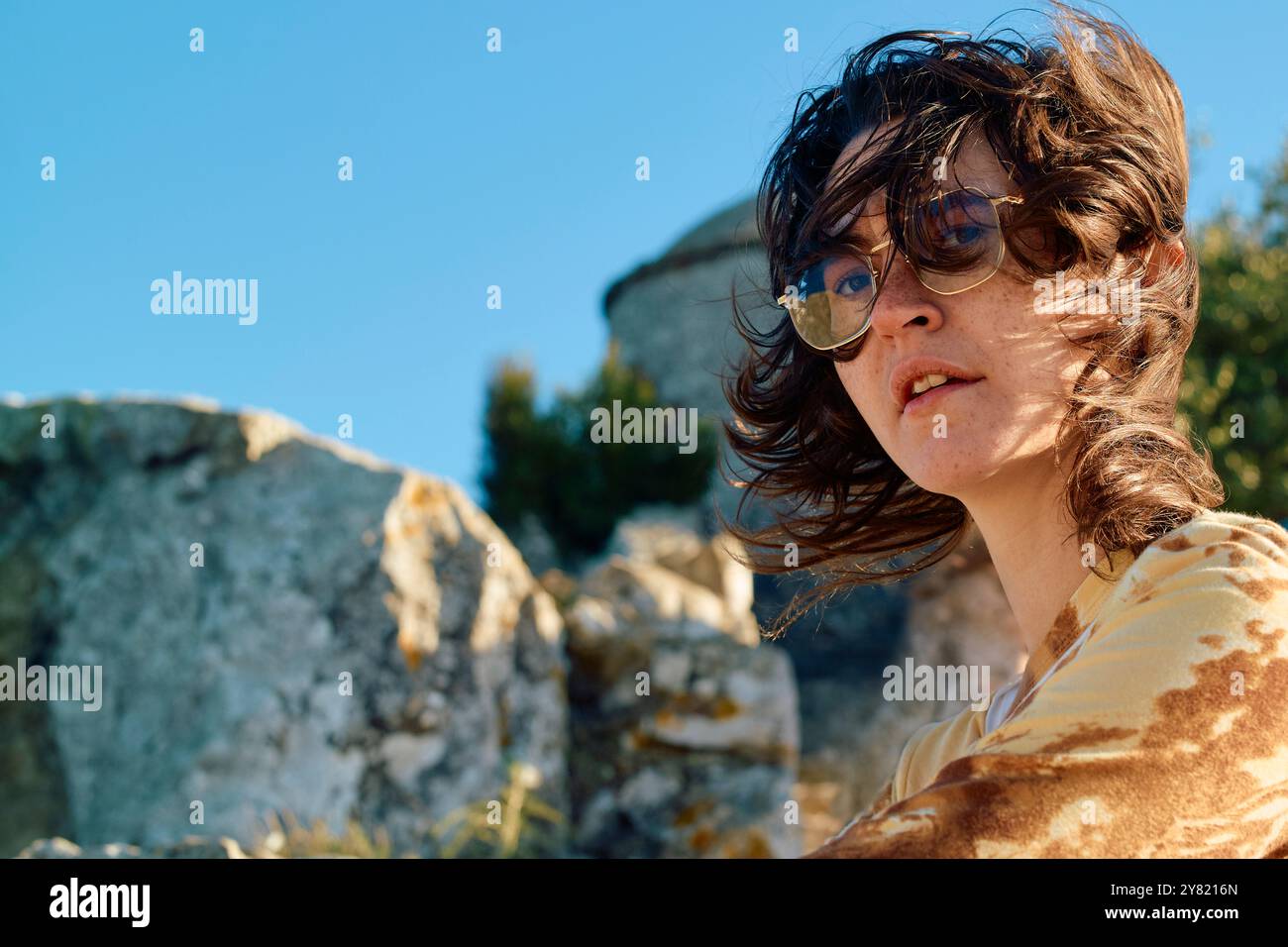 Windgepeitschte Frau mit gewelltem Haar und Brille draußen mit alten Steinstrukturen im Hintergrund. Stockfoto