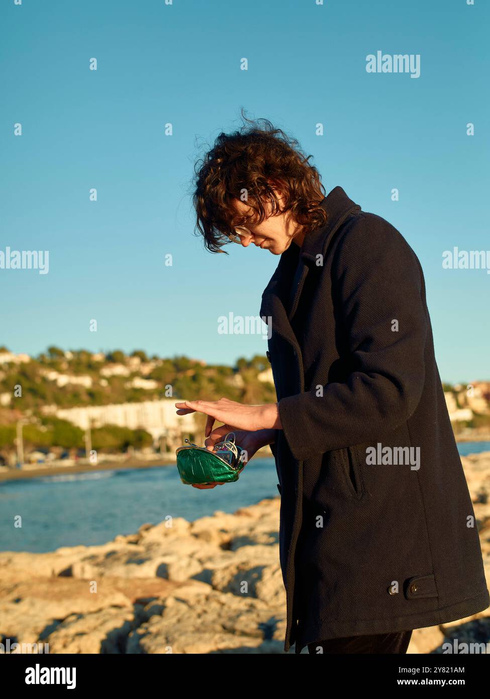 Lockenhaarige in dunklem Mantel steht am Meer und gießt Flüssigkeit aus einer grünen Flasche. Stockfoto