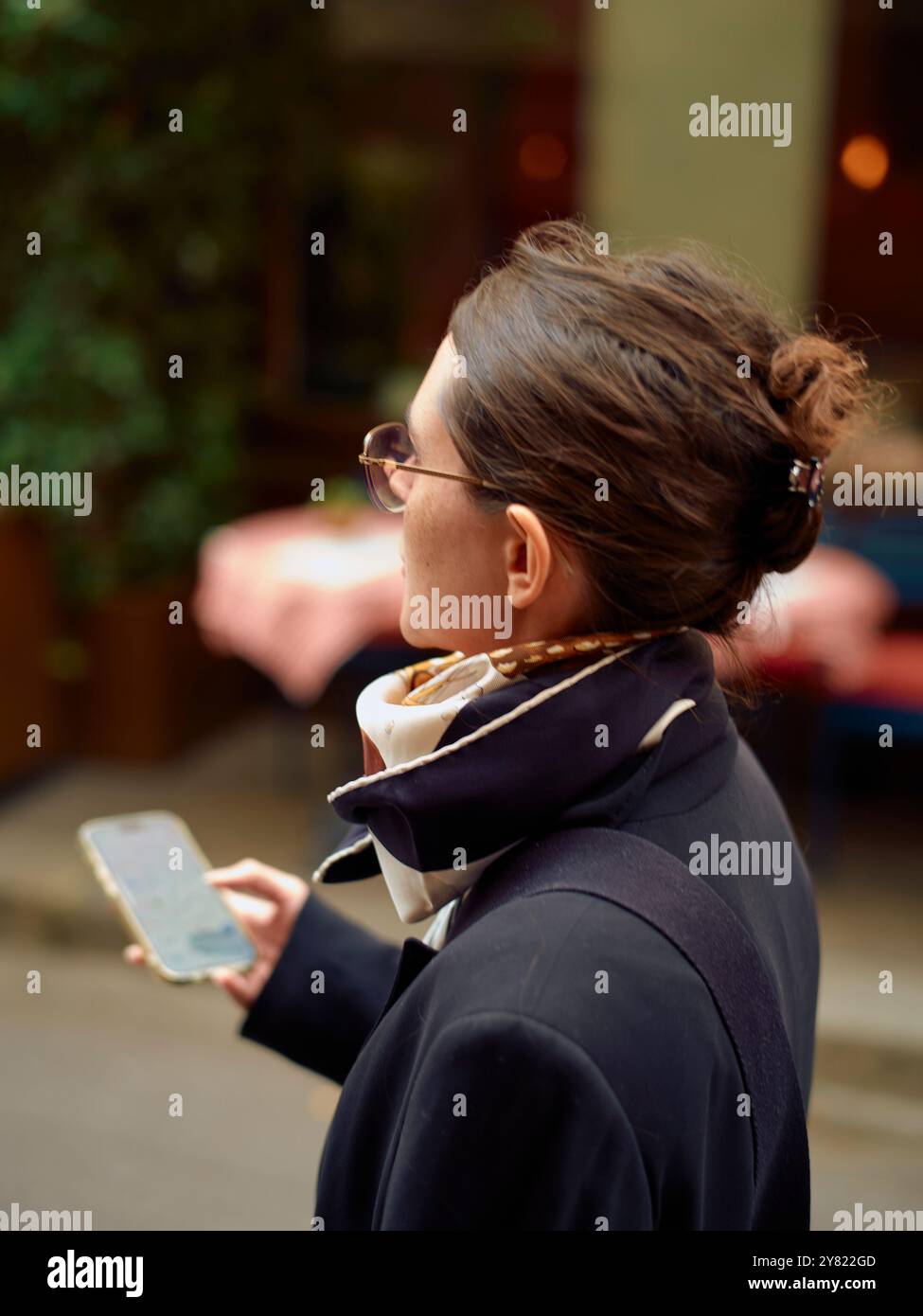 Eine Frau in Brille hält ein Smartphone, vertieft in das Lesen des Bildschirms, während sie draußen steht. Stockfoto