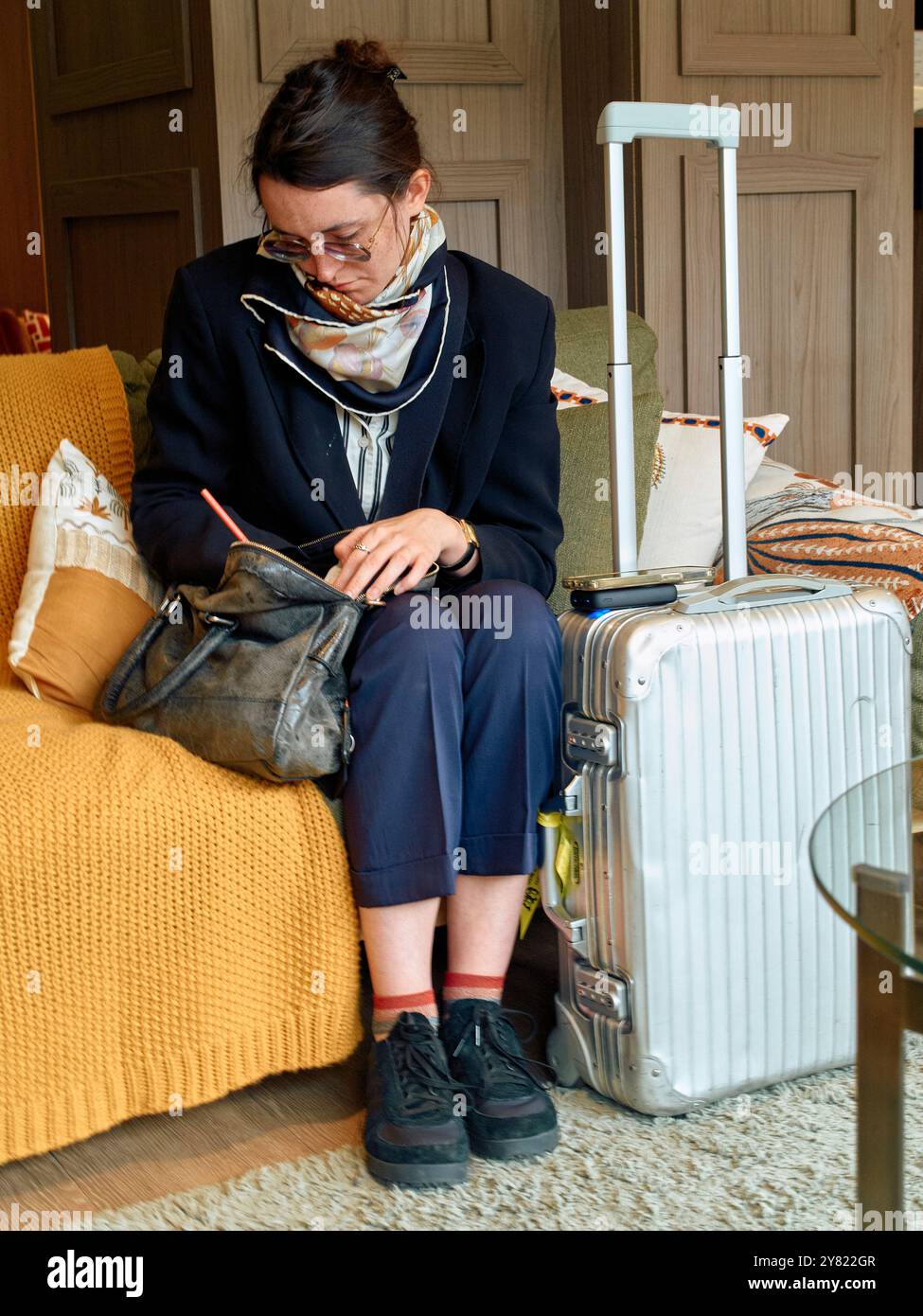 Frau, die auf einer gelben Couch sitzt und ihr Handy mit einem weißen Koffer und einer braunen Tasche neben ihr ansieht. Stockfoto
