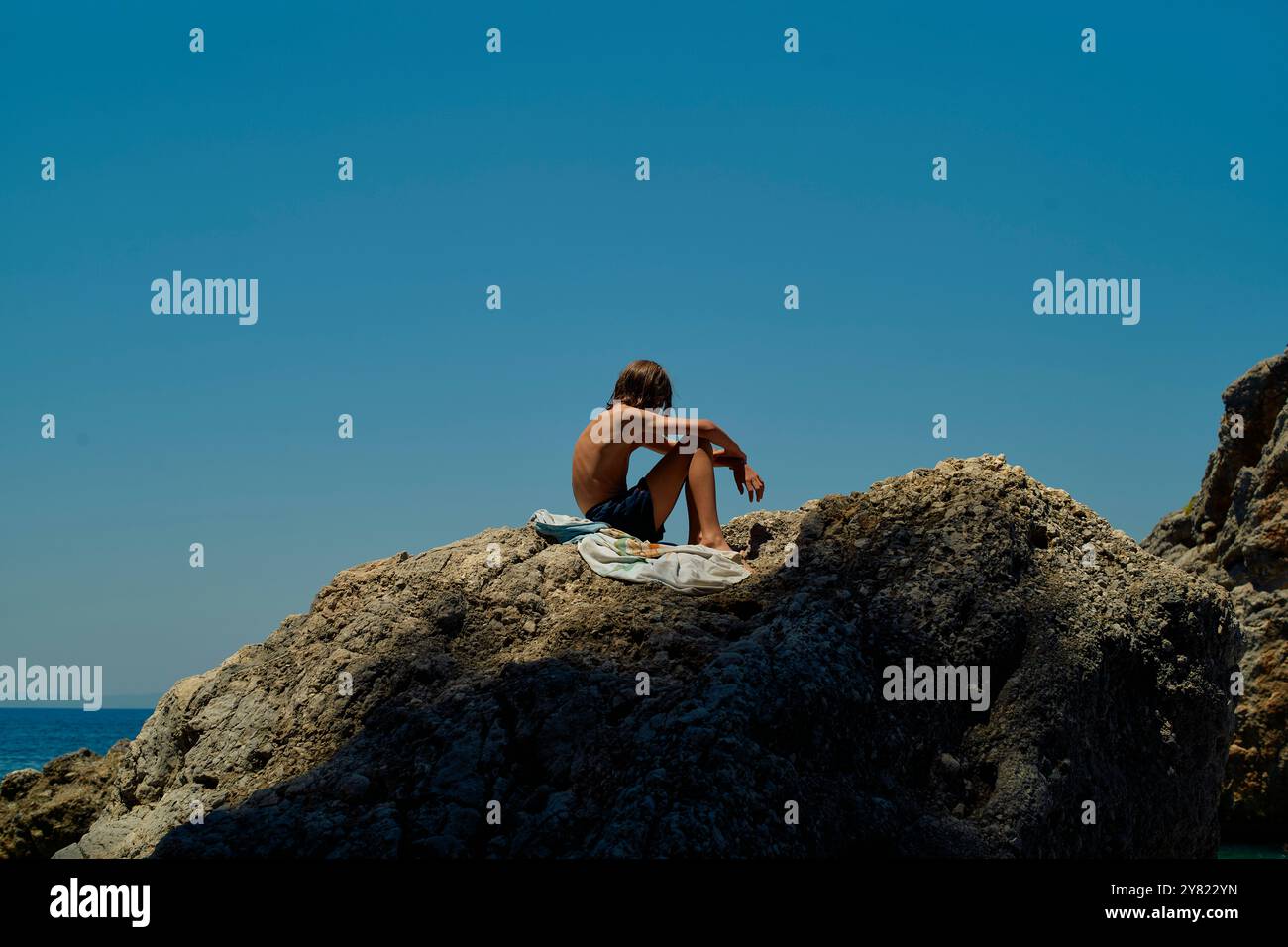 Eine Person, die auf einem Felsvorsprung am Meer sitzt und an einem hellen sonnigen Tag den Horizont betrachtet. Stockfoto