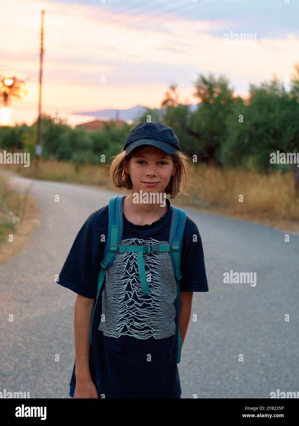 Ein lächelnder Teenager mit Rucksack steht bei Sonnenuntergang auf einer Landstraße mit blauem Hut und T-Shirt. Stockfoto