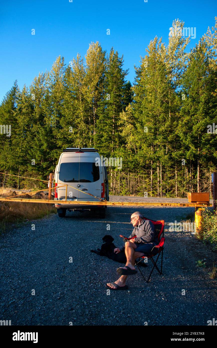 Ein älterer Mann, der sich an einer Straßensperre entspannt, ein Kindle-Buch liest und sein Schwarzes Labor streichelt. Mount Rainer National Park, Washington Stockfoto