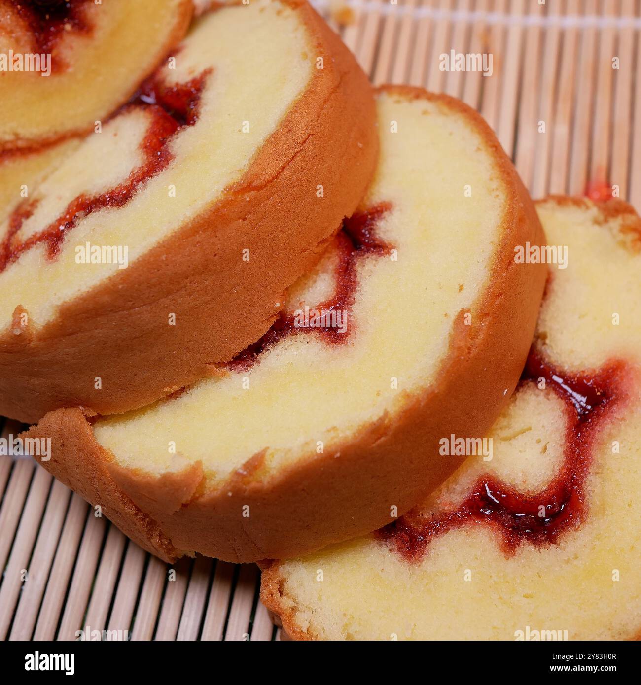 Gerollte Kuchenscheiben auf Bambusmatte. Nahaufnahme mehrerer gerollter Kuchen mit roter Marmelade im Inneren, angeordnet auf einem Bambusständer. Stockfoto