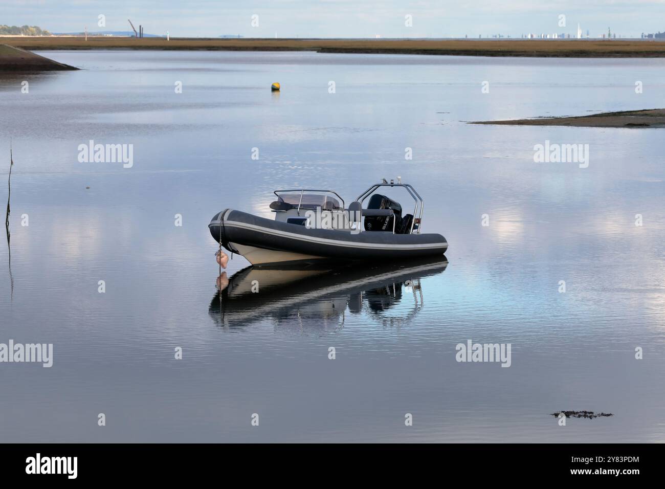 Ein Motorboot, das im stillen Wasser verankert ist, dessen Reflexion sichtbar ist, mit Land im Hintergrund. Stockfoto