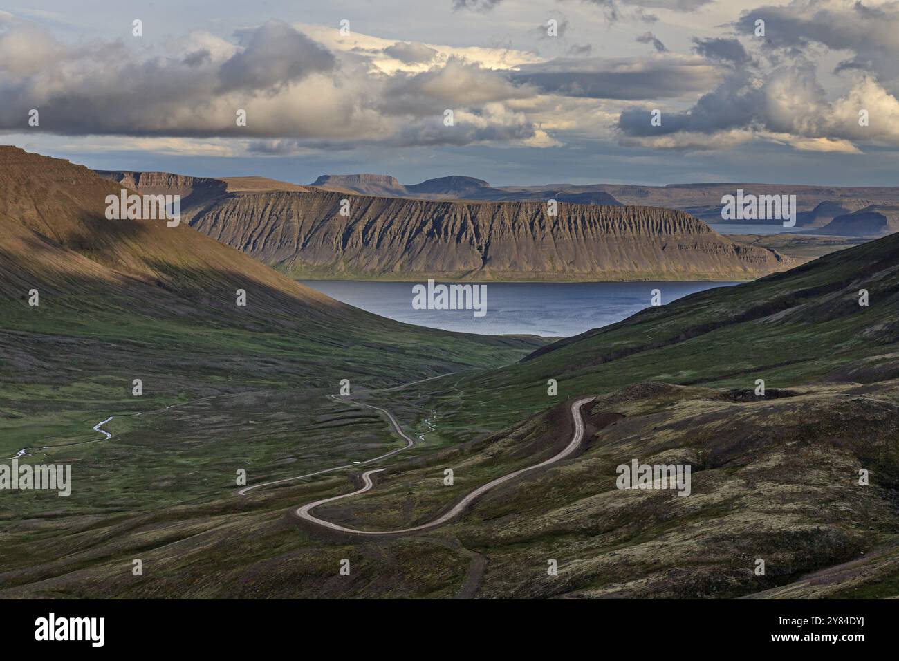 Gewundene Schotterstraße, Bergstraße, Fjord und Berge dahinter, bewölkte Stimmung, Sommer, Westfjorde, Island, Europa Stockfoto