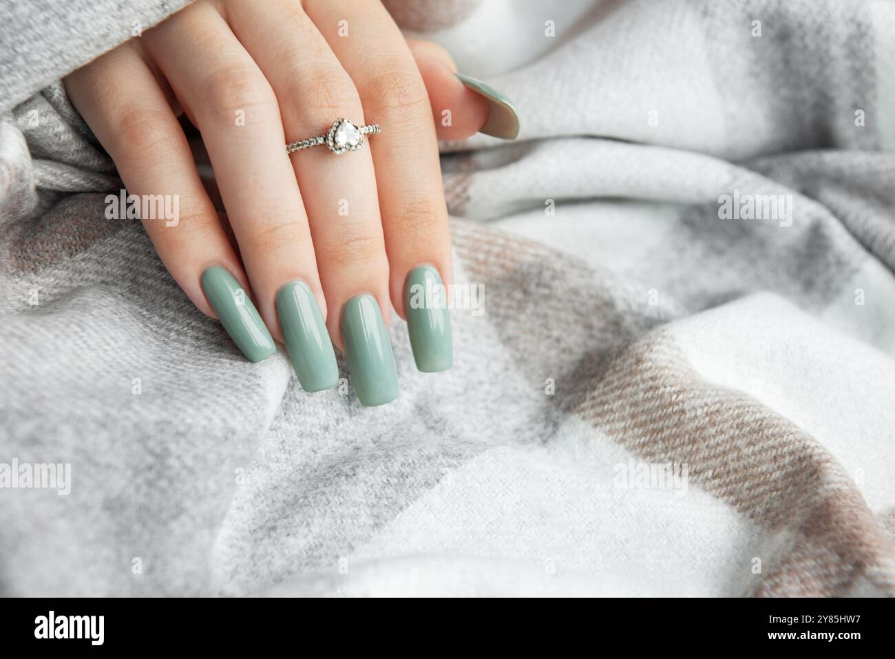 Eine Hand zeigt lange, elegant gepflegte Nägel, die in einem weichen Grünton lackiert sind. Die Hand liegt auf einer gemütlichen, gemusterten Decke, die ein funkelndes Licht hervorhebt Stockfoto