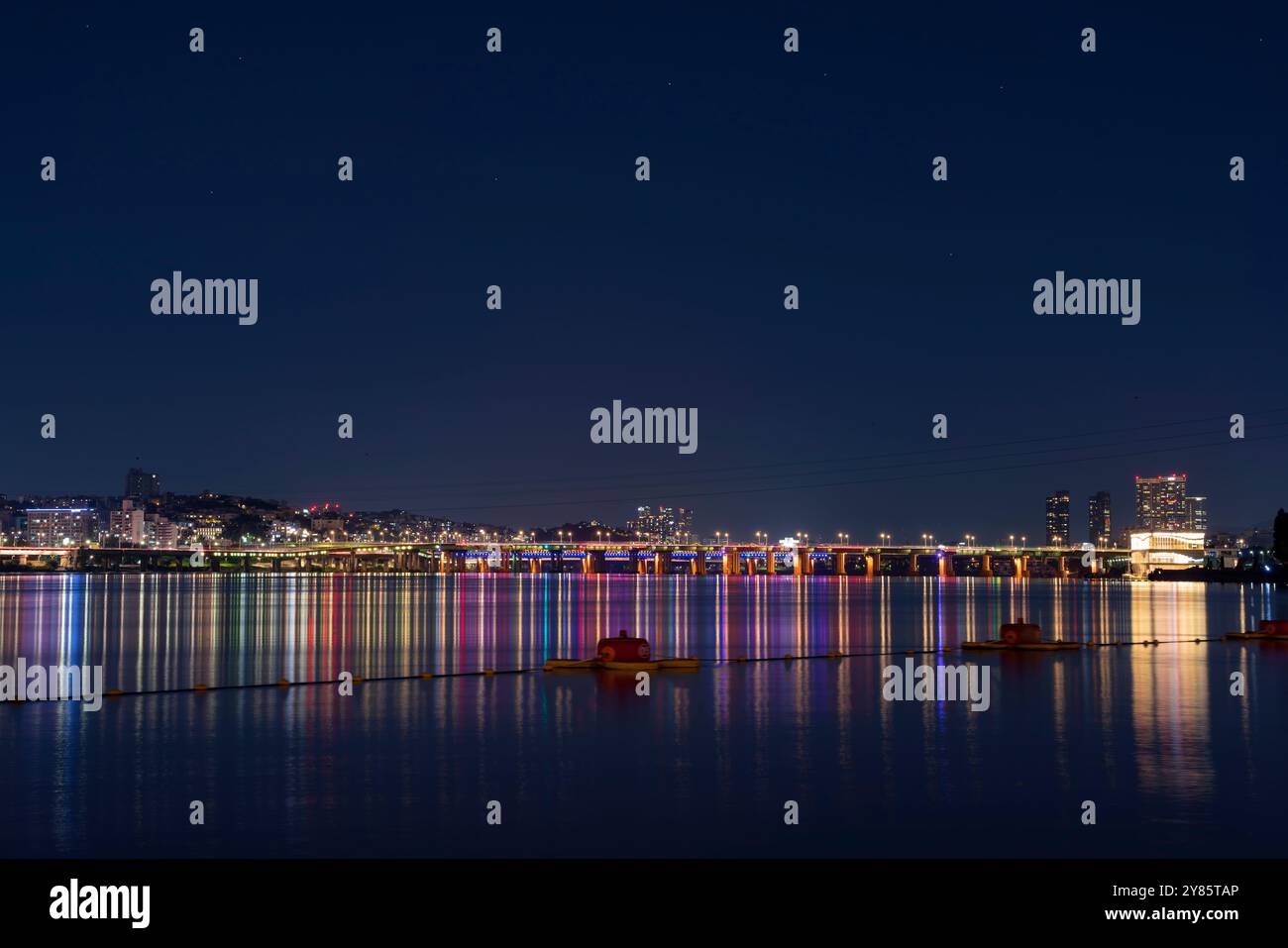 Dieses Foto zeigt einen nächtlichen Blick auf einen Park, in dem die Lichter der Stadt auf dem ruhigen Flusswasser reflektieren und die beleuchtete Brücke die ruhige Atmosphäre unterstreicht. Stockfoto