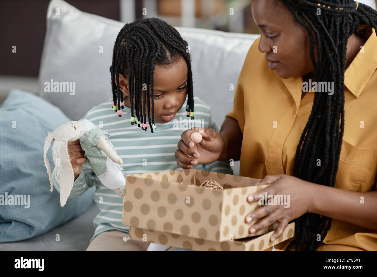 Afroamerikanische Mutter und Tochter halten die Puppe, sitzen auf der Couch, während sie das Präsent mit einer gestrichelten Schachtel auspacken. Sie zeigen Neugier und Engagement im Geschenk Stockfoto