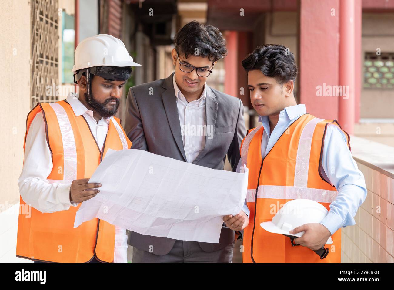 Ein Architekt, Bauingenieur und Innenarchitekt untersucht eine große Reihe von Bauplänen auf einer geschäftigen Baustelle und diskutiert sie. Stockfoto