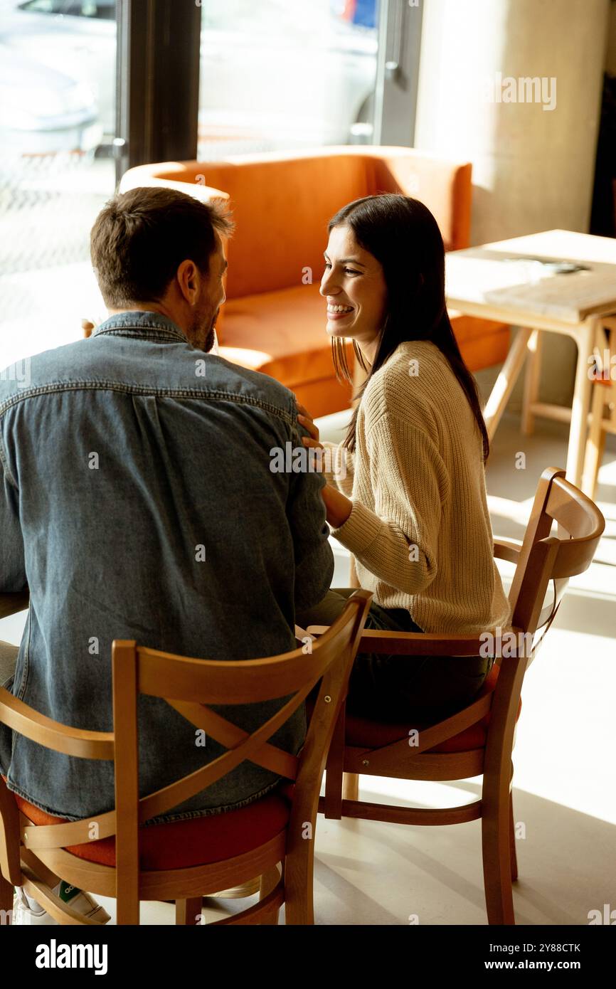 Ein Paar teilt sich einen freudigen Moment in einem Café und schlürft erfrischende Getränke, während er an einem Holztisch sitzt, umgeben von warmem Sonnenlicht und einem einladenden atmos Stockfoto