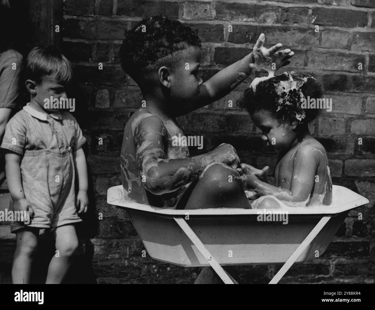 Obwohl ich es selbst sage, weiß ich nicht viel über Frisuren. Mama wird dich einfach nicht erkennen, wenn ich fertig bin. Michael Agbadabunia, 4 Jahre alt, und seine achtzehn Monate alte Schwester Sandra hatten in ihrem Haus in der Radnor-Street, Hulme, Manchester eine Badewanne im Freien. Abkühlung. Während Sydneysider zittern, hat England Hitzewellen erlebt. Letzte Woche in Manchester kühlten sich die beiden Jugendlichen mit einem Freiluftbad ab. Juli 1952. Stockfoto