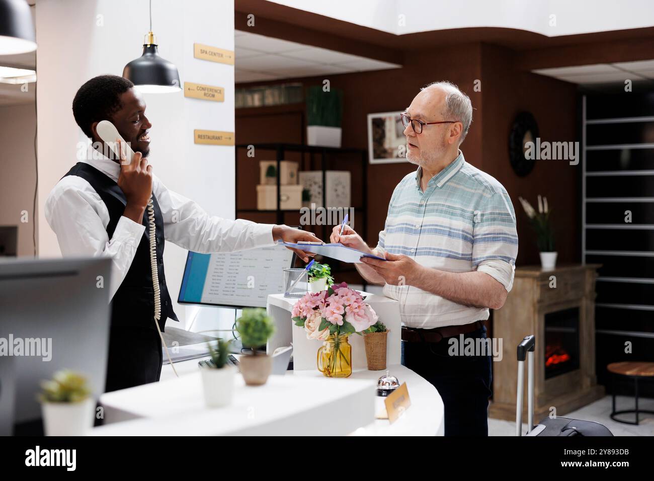Ein älterer kaukasischer Mann im Ruhestand unterschreibt Reservierungsunterlagen am Schalter der Hotellobby, während ein afroamerikanischer Angestellter einen Anruf entgegennimmt. Der leitende Kunde füllt die Checkin-Papiere aus, da der Rezeptionist am Telefon ist. Stockfoto