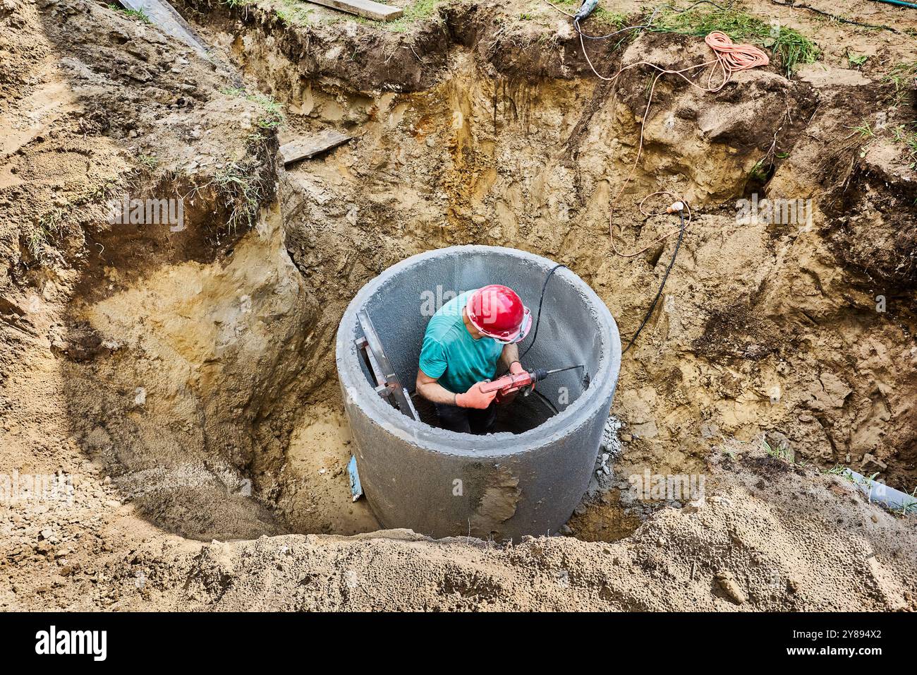 Der Klärgrubenbauer zerquetscht eine Wand aus Betonring mit Bohrschnur bei der Installation einer autonomen Abwasseranlage in einem privaten Landhaus. Stockfoto