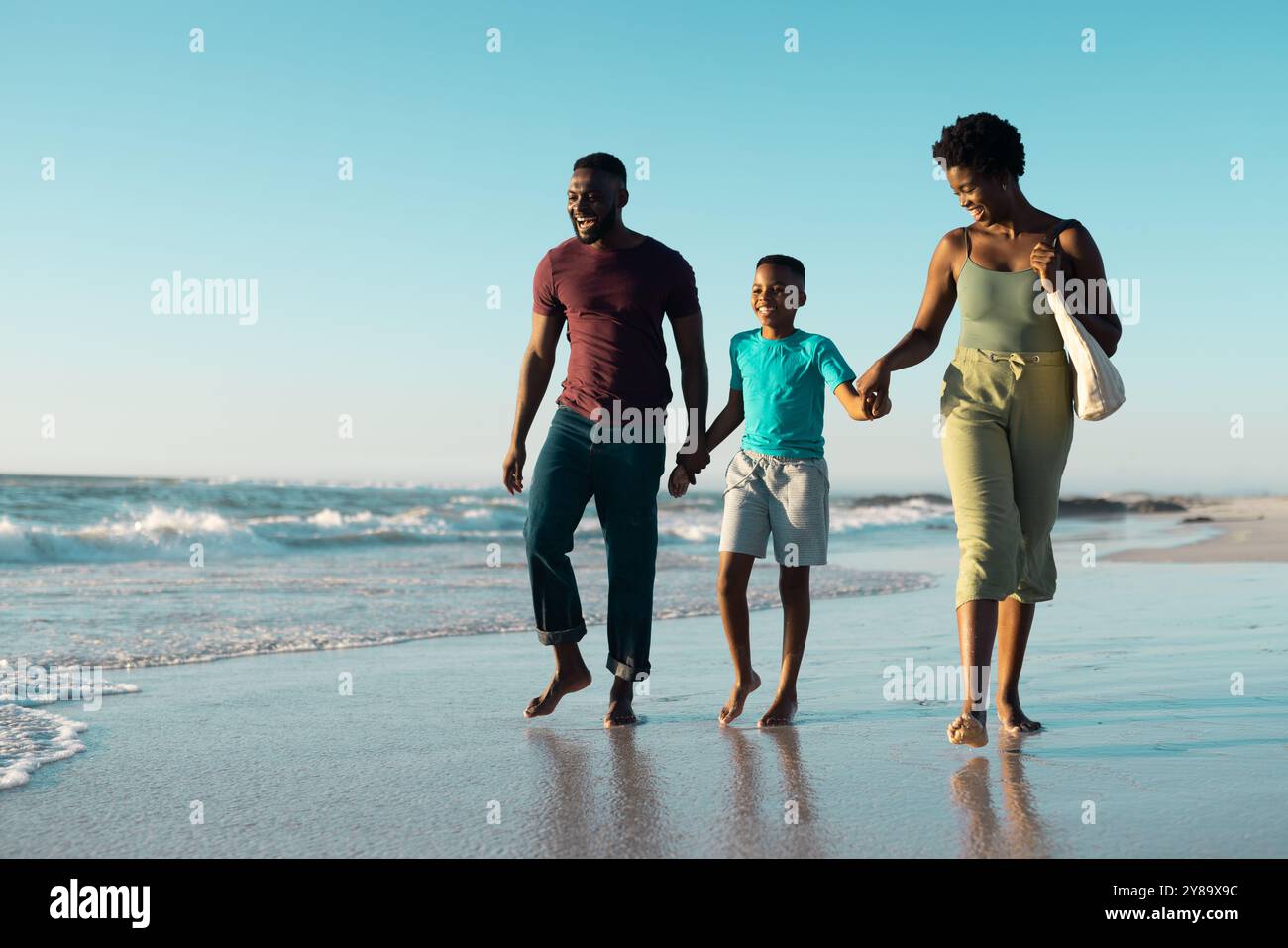 Fröhlicher afroamerikanischer Junge, der Vater- und Mutterhände hält und am Ufer gegen den Himmel läuft Stockfoto