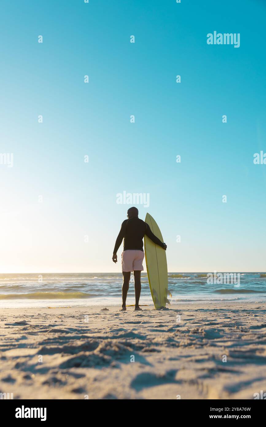 Rückansicht eines afroamerikanischen Seniorenmannes mit Surfbrett, der das Meer und den klaren Himmel während des Sonnenuntergangs betrachtet Stockfoto