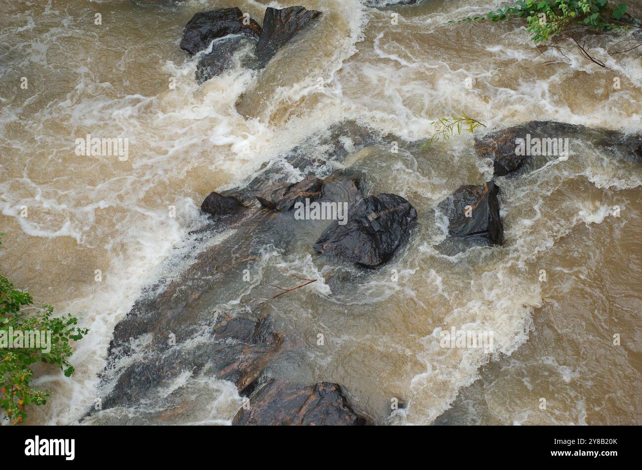 Close Up Overhead Blick hinunter auf die Flussfelsen in einem schnell bewegenden, schlammigen, braunen Fluss, der mit weißen Kappen rollte. Aggresse Stockfoto
