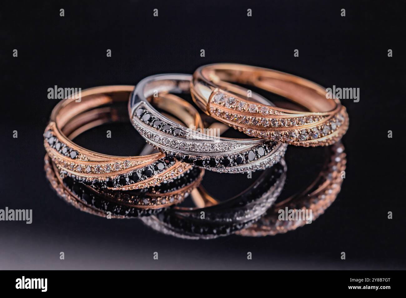 Eheringe werden im Spiegel reflektiert. Ein Satz von drei Ringen auf schwarzem Hintergrund. Gold- und Silberringe mit schwarzen und weißen Steinen. Stockfoto
