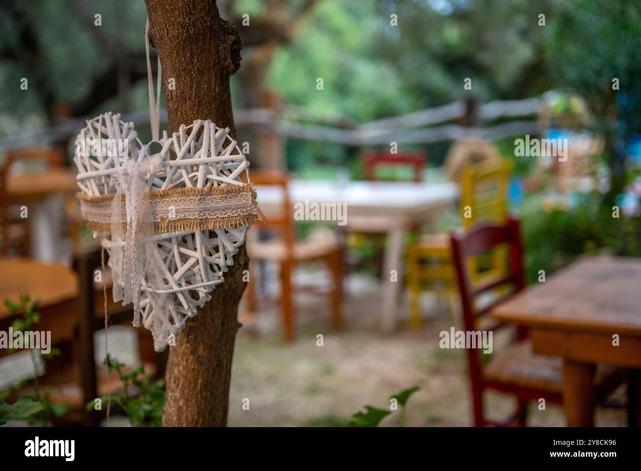 Handgefertigtes Strohherz, das an einem Baum als Dekoration in einer griechischen Taverne mit Tischen und Stühlen im Hintergrund hängt Stockfoto