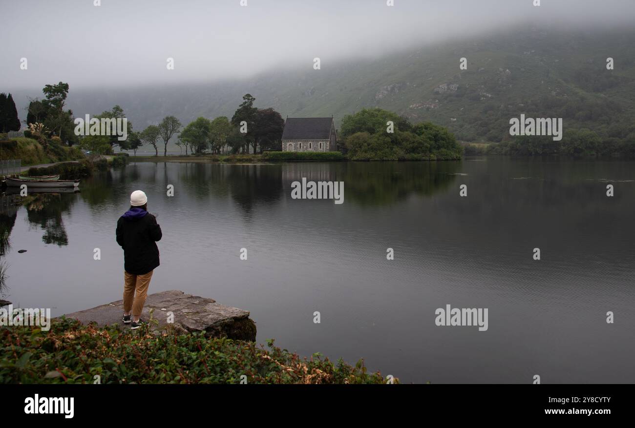 Touristen bewundern die malerische Aussicht auf eine Kirche auf einem See in irland an einem nebeligen Tag. Gougane barra Chuch Stockfoto