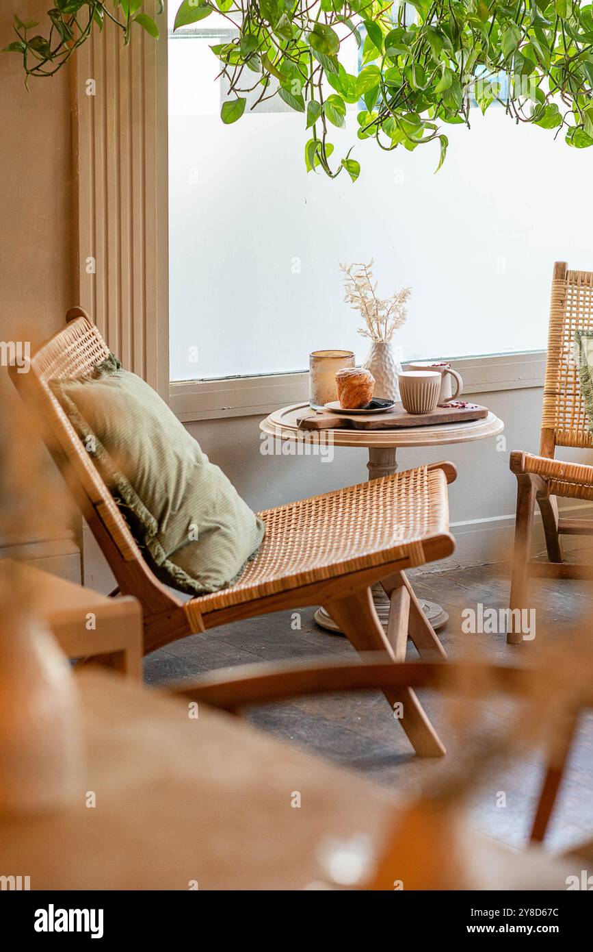 Gemütliche Café-Ecke mit Rattanstuhl, kleinem runden Tisch mit Kaffee, Gebäck und einer Vase mit getrockneten Blumen, umgeben von üppigen hängenden Pflanzen Stockfoto