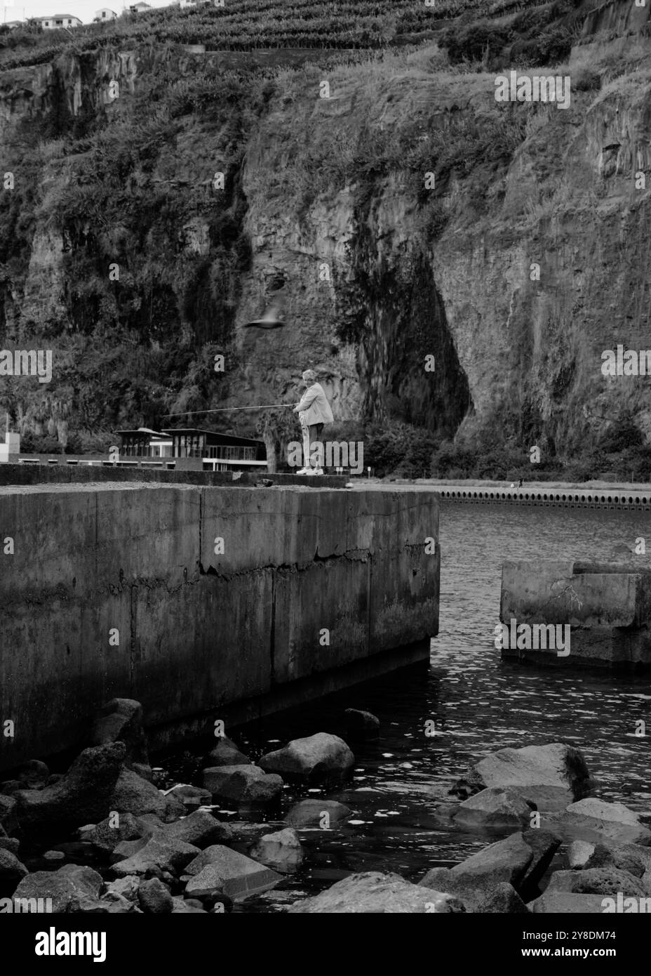 Monochrom einer einsamen Fischerin, die auf einem zerklüfteten Betonpier mit hohen Klippen entlang der Küste Madeiras steht Stockfoto