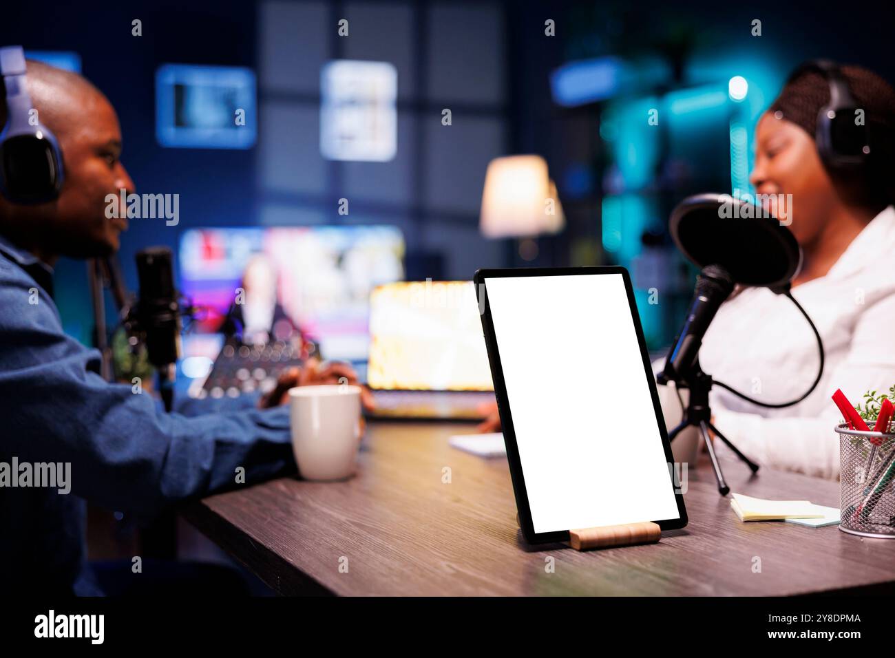 Männliche und weibliche Inhaltsersteller, die Kopfhörer tragen, streamen live, während das Tablet einen leeren Bildschirm anzeigt. Schwarze Podcaster, die Geräte auf dem Schreibtisch verwenden, während das Gerät eine Kopierraum-Modellvorlage anzeigt. Stockfoto