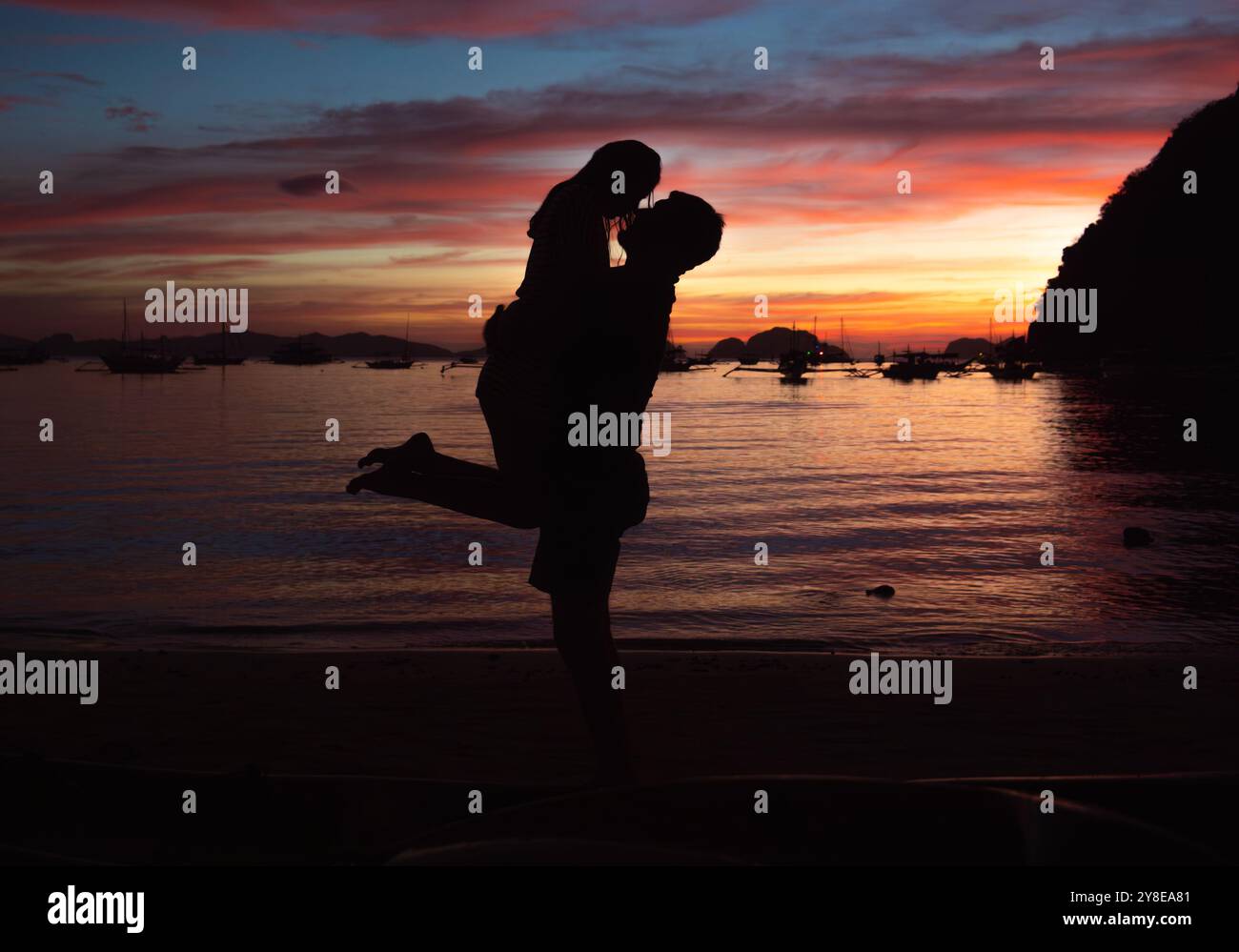 Ein Paar umarmt sich fröhlich, in Silhouette gefangen, während der Himmel bei einem wunderschönen Sonnenuntergang an einem ruhigen Strand mit leuchtenden Farben gemalt wird. Stockfoto