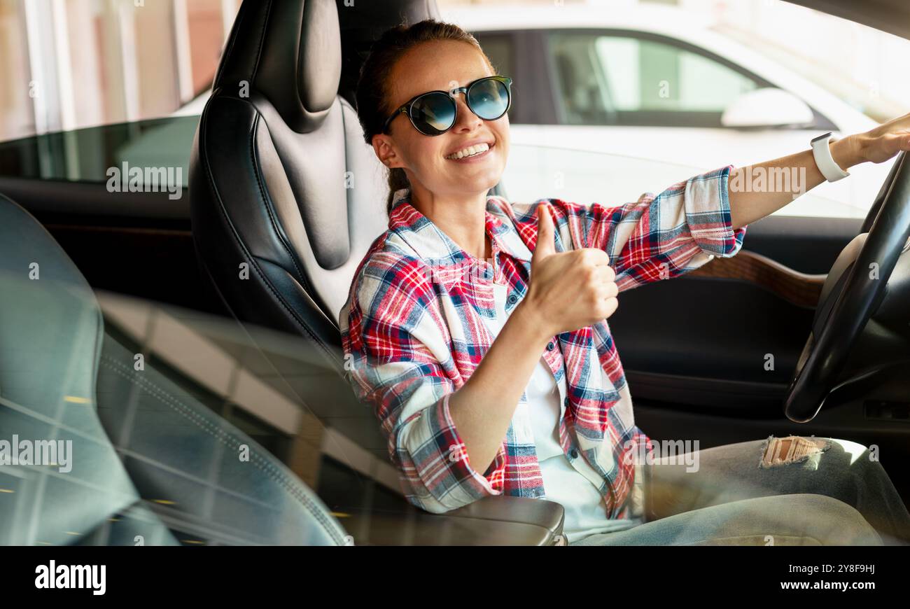 Eine junge Frau sitzt in ihrem neuen Auto im Händlerbetrieb, lächelt und gibt ein Schild mit dem Daumen nach oben. Autovermietung, Carsharing. Stockfoto
