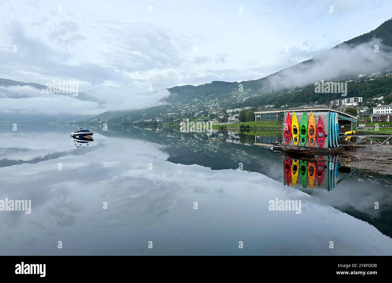 Farbenfrohe Kajaks spiegeln die Reflexion auf dem friedlichen Bergsee am nebeligen Morgen, Schneestücken auf dem Hügel auf der linken Seite. Stockfoto