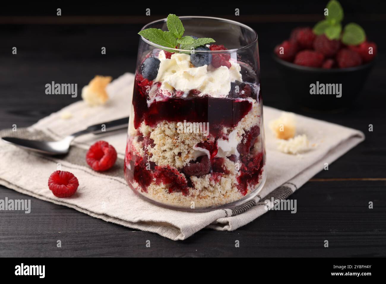 Leckeres kleines Dessert. Beeren, Biskuitkuchen und Schlagsahne im Glas auf schwarzem Holztisch Stockfoto