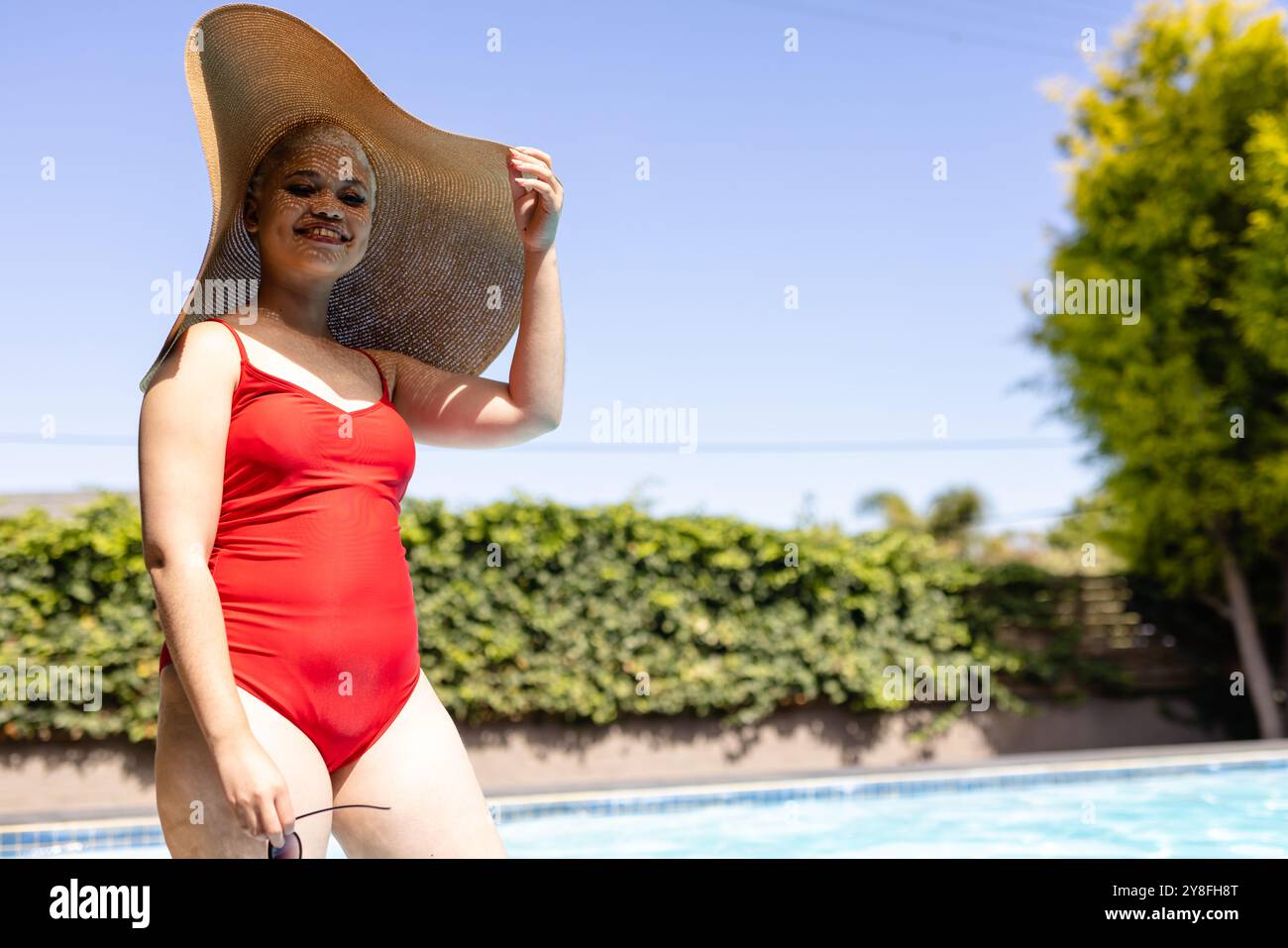Glückliche Frau in Badeanzug und Sonnenhut am Pool, lächelnd im Sonnenlicht, Kopierraum Stockfoto