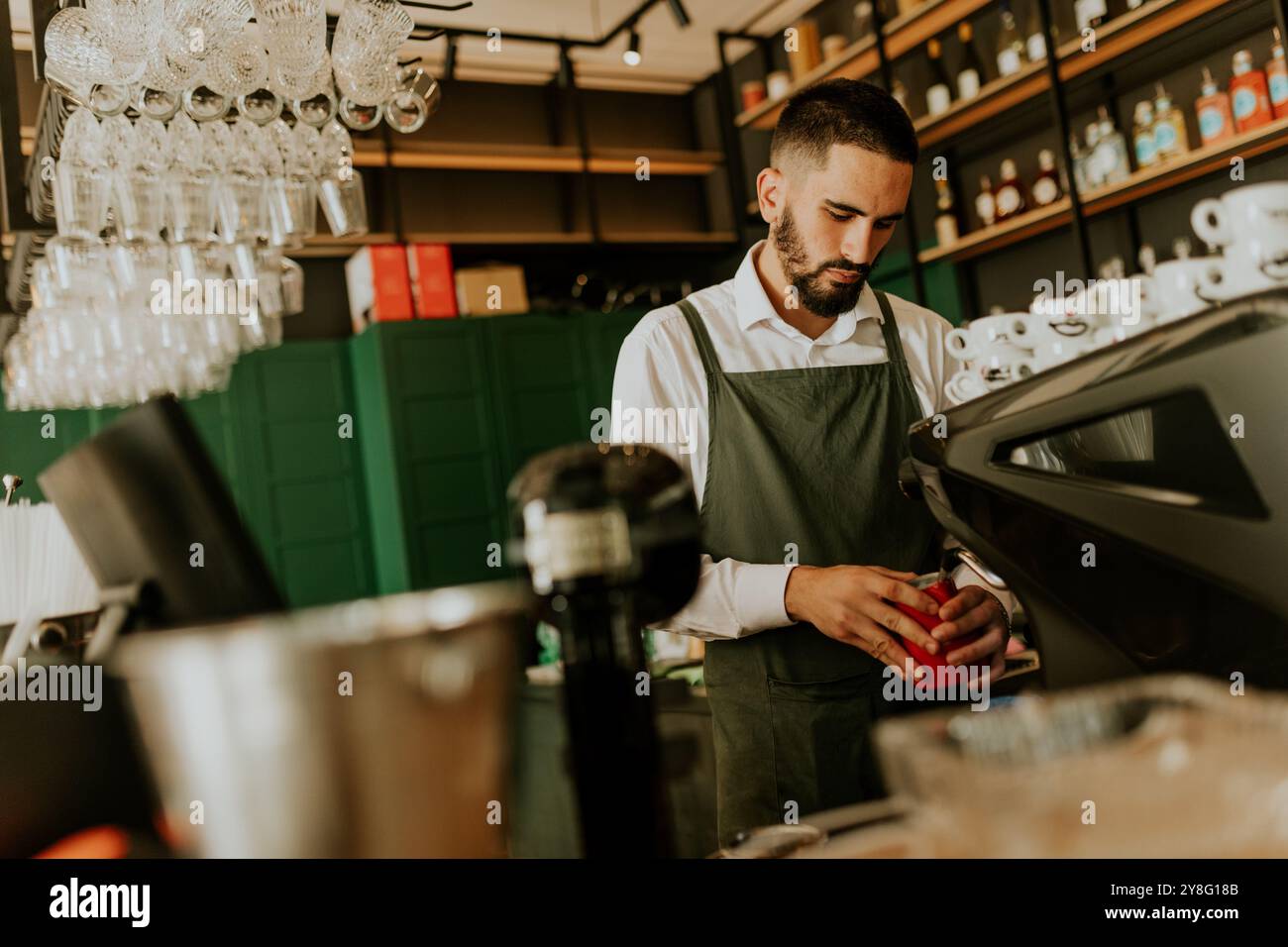 In einem schicken Café konzentriert sich ein erfahrener Barista auf die Herstellung köstlicher Getränke, umgeben von ordentlich angeordneten Gläsern und lebendiger Einrichtung, die eine warme Atmosphäre schaffen Stockfoto